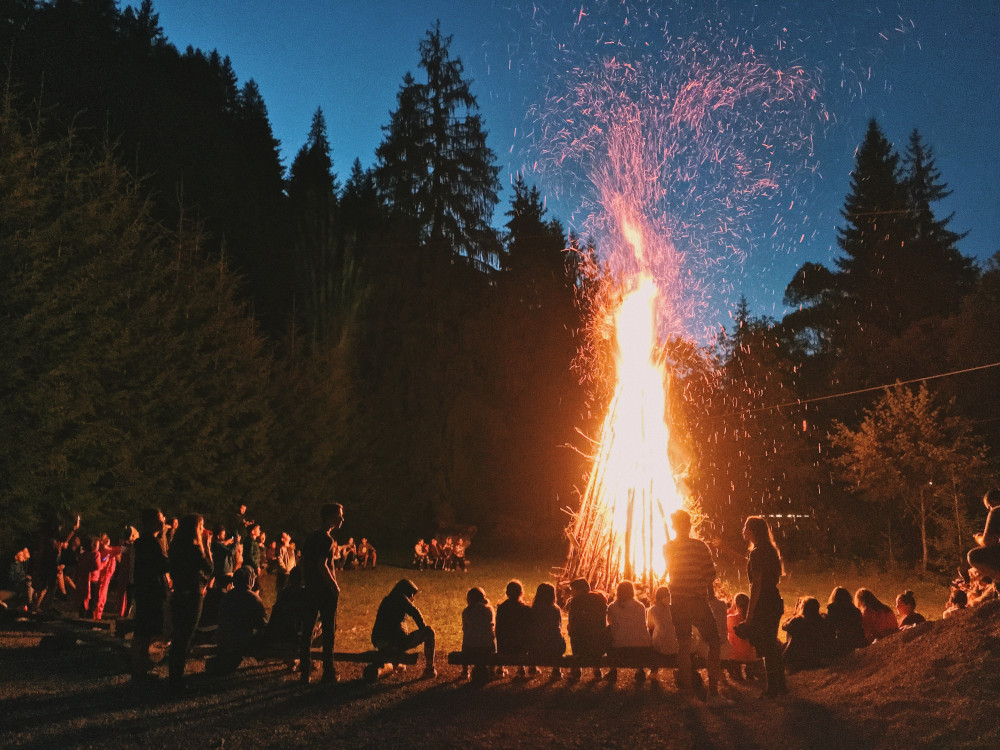 a group of young people stand and sit on benches watching a bonfire that reaches more than twenty feet in the air