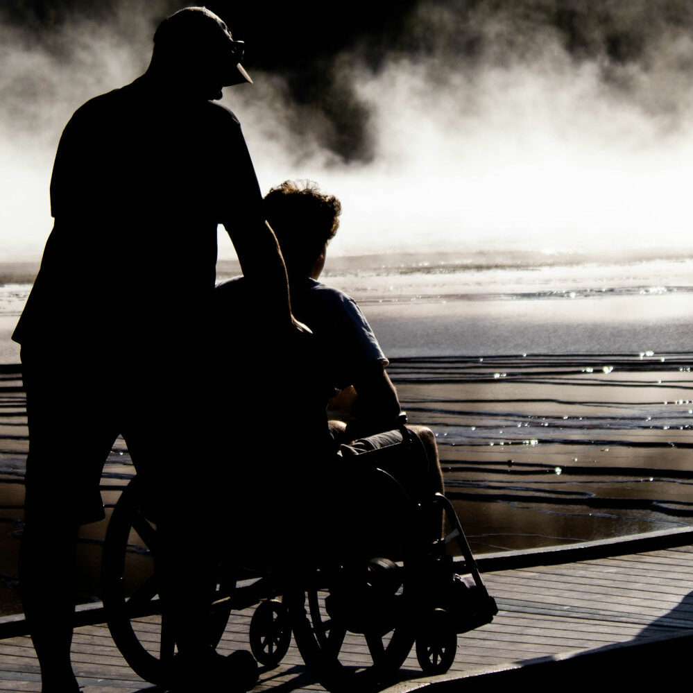 Silhouette of a man pushing someone in a wheelchair across a boardwalk ramp