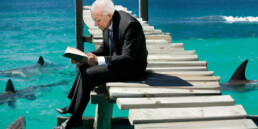 old man in a suit sitting on a dock reading to sharks